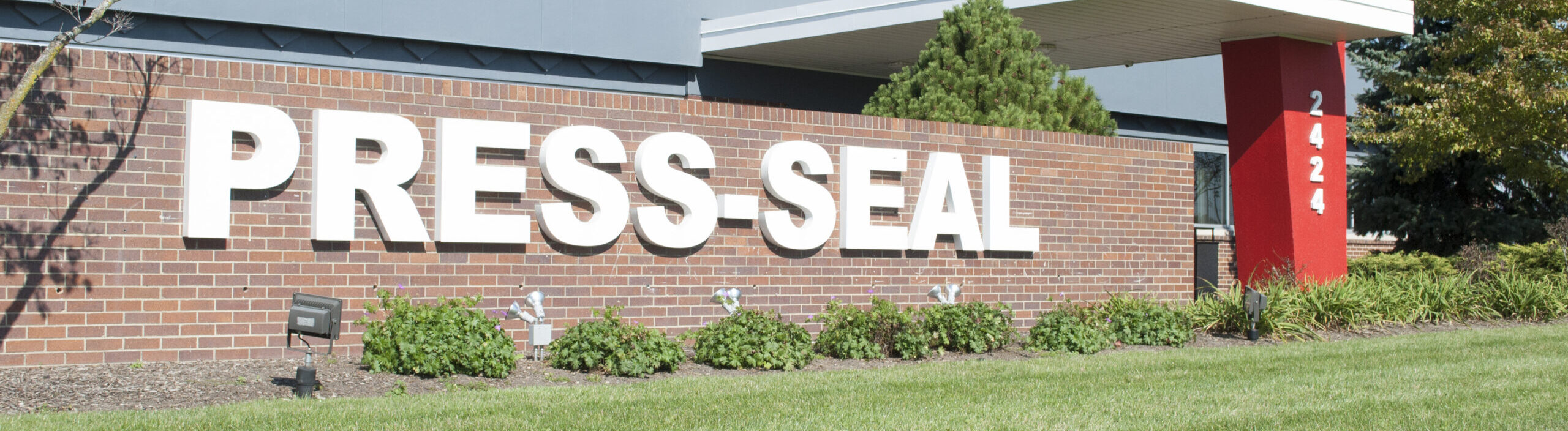 Grassy photo with brick wall in background and blue skies representing a manufacturing building
